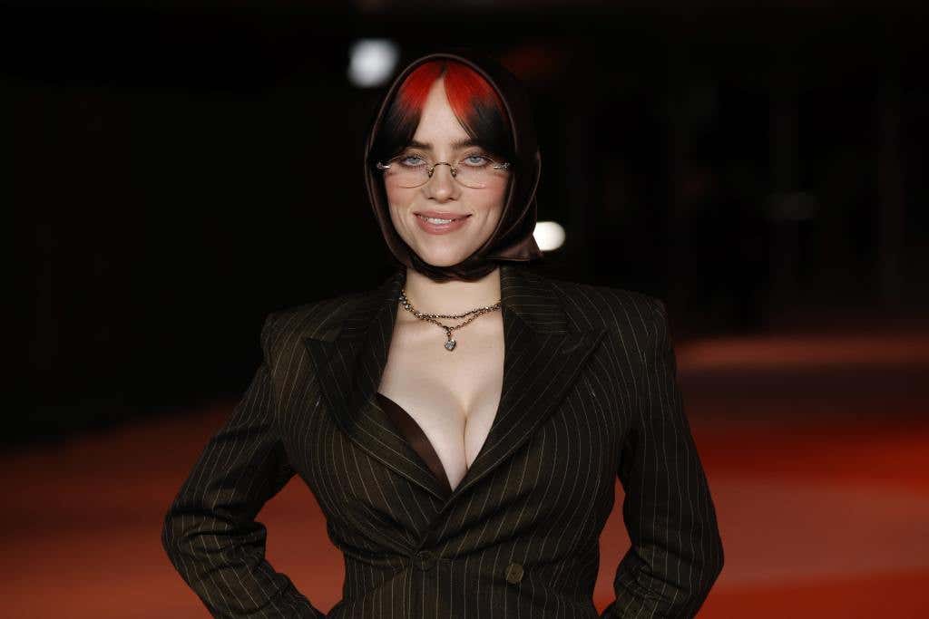 Billie Eilish attends the 3rd Annual Academy Museum Gala smirking wearing a brown silk head wrap, clear glasses, and pin stripe blazer.