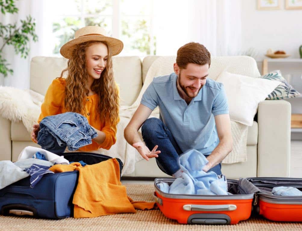 Laughing man and woman in summer hat setting off for journey and packing bags on floor in living room anticipating vacation, pre-vacation prep concept