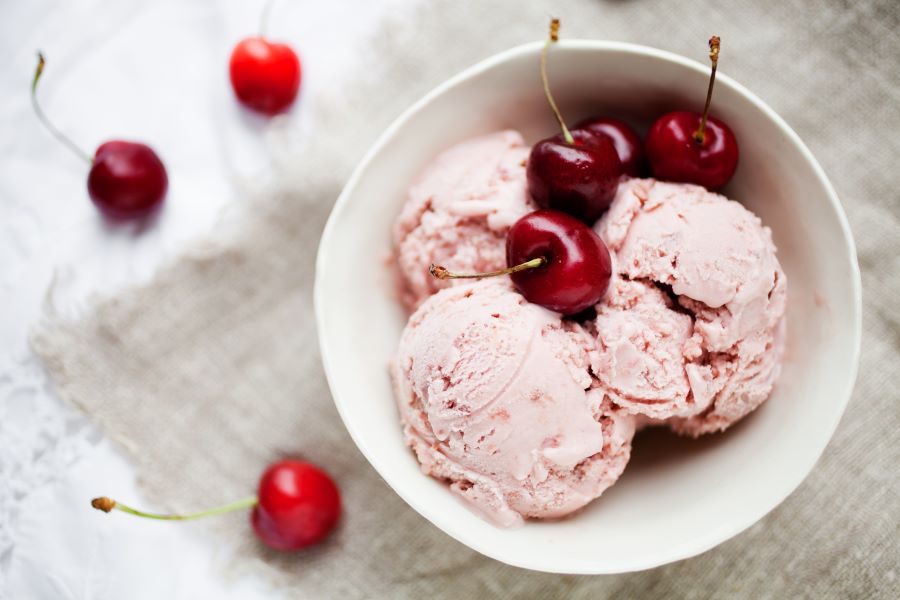 White bowl with three scoops of pink ice cream topped with red cherries with stems, a colorful 4th of July dessert